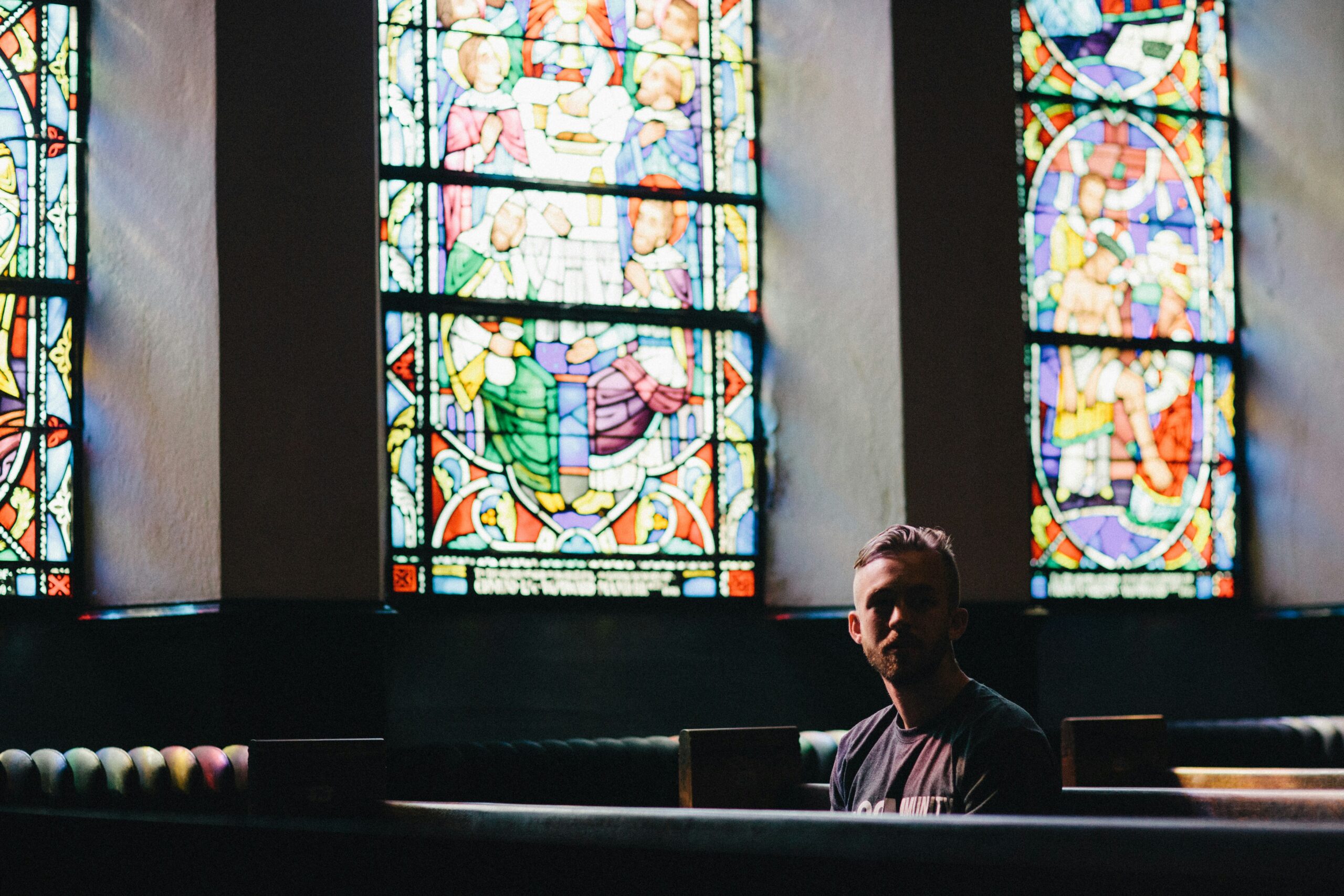 A person sitting in a dimly lit church pew with colorful stained glass windows in the background, depicting religious scenes. The light from the windows casts vibrant patterns on the pews.