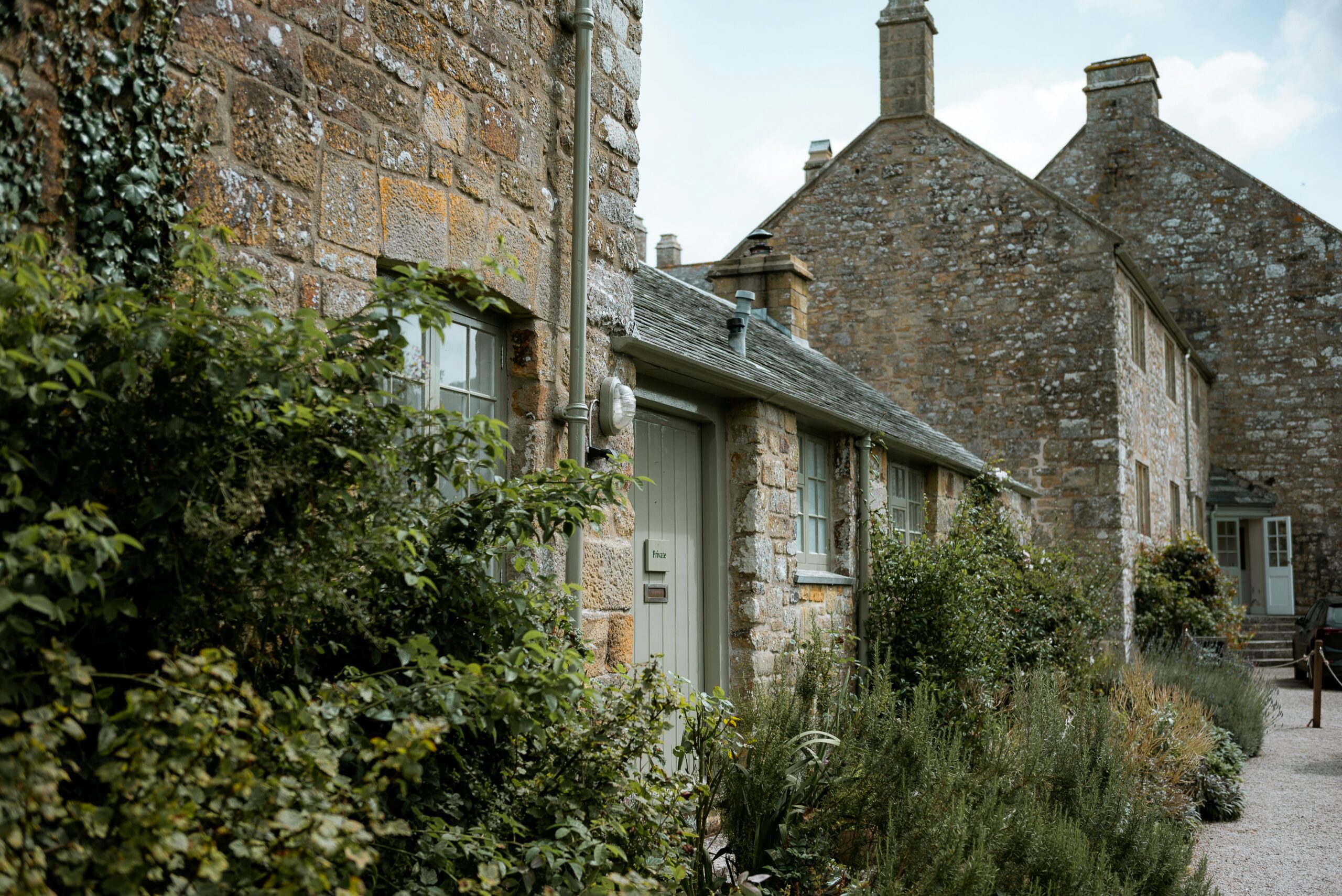 A row of charming stone cottages with ivy and greenery growing around them. The buildings have slate roofs and small, multi-paned windows. A gravel path runs alongside the cottages, bordered by lush plants and shrubs.